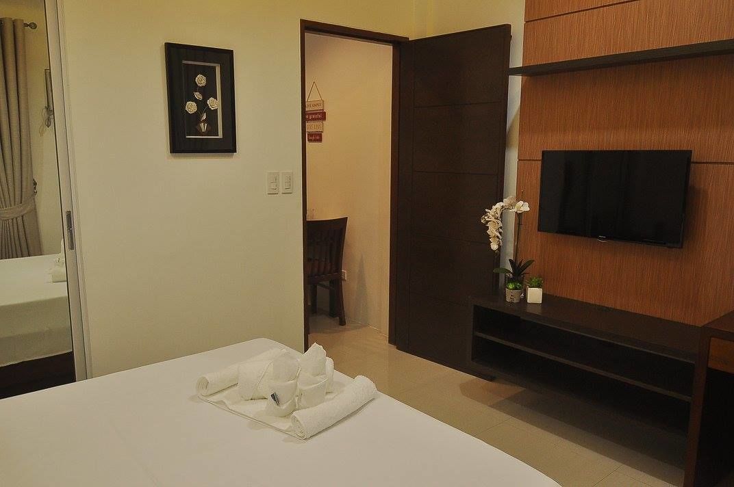 A hotel room with a bed, a TV mounted on the wall, a door leading to another room, and a small table with a vase of flowers.