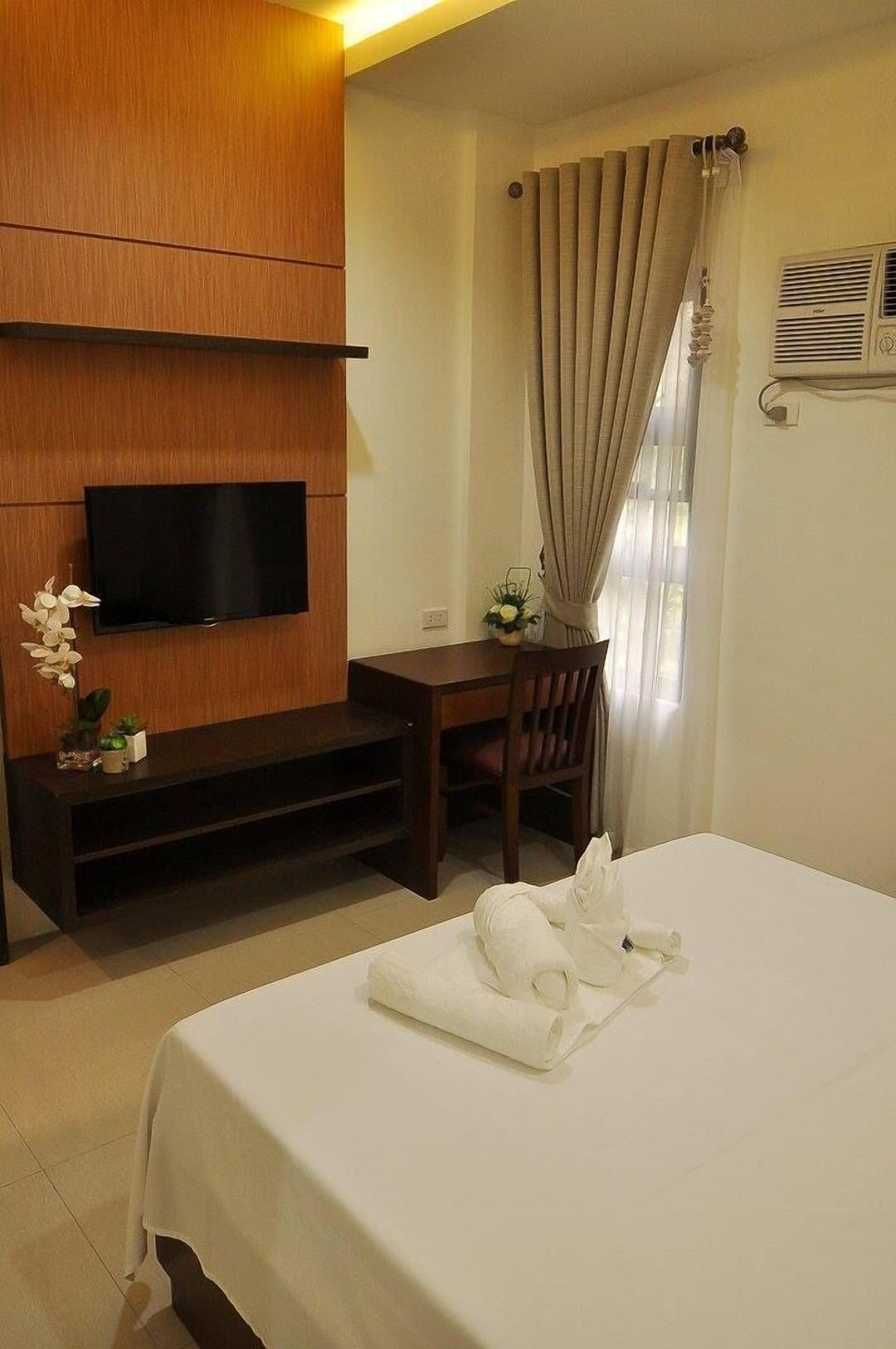 A hotel room with a bed, a TV mounted on a wooden panel, a desk with a chair, and a window with curtains.