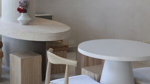 A modern dining area with a round table and a chair, a white vase with flowers, and a shelf with decorative items.