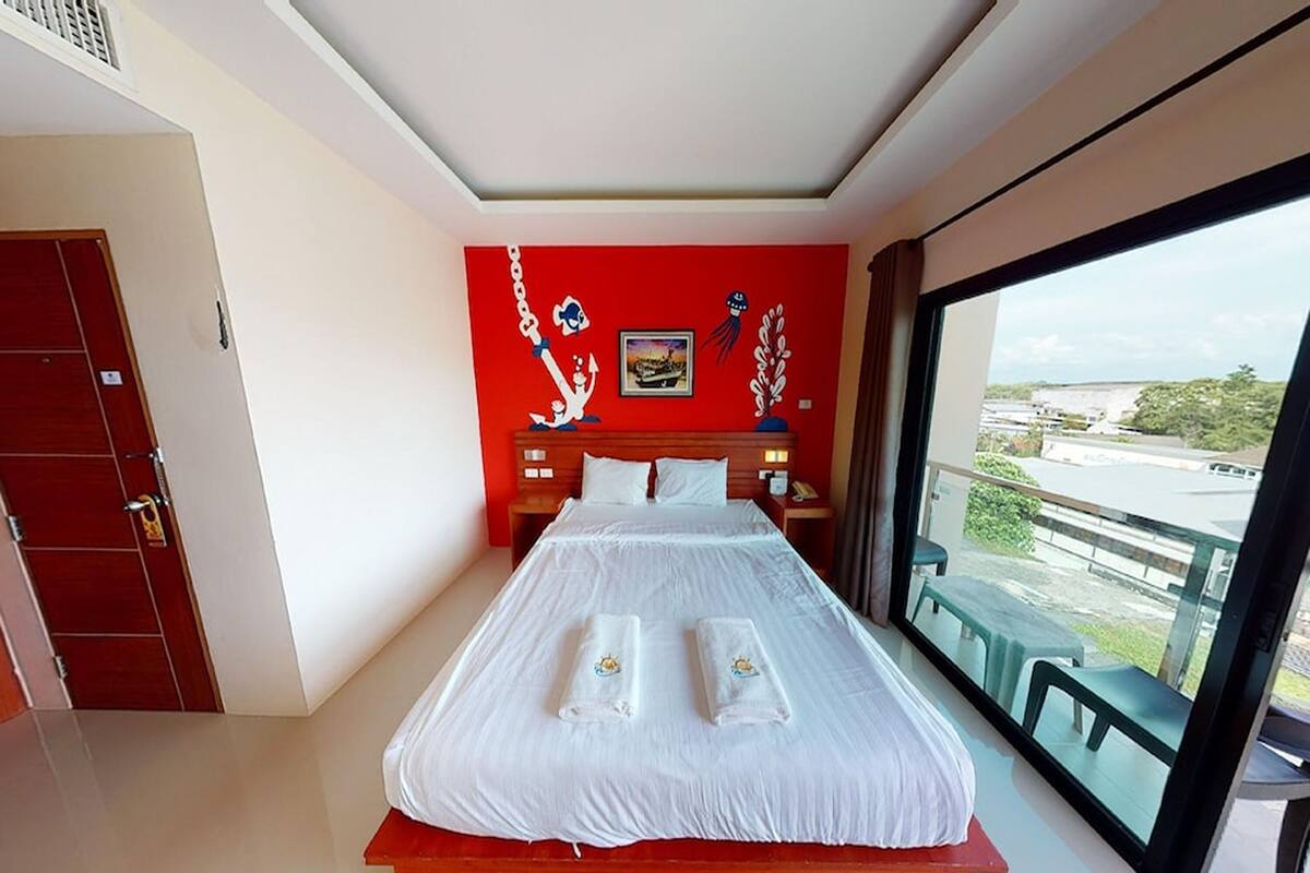 A hotel room with a large bed, a red accent wall, and a balcony with a view.