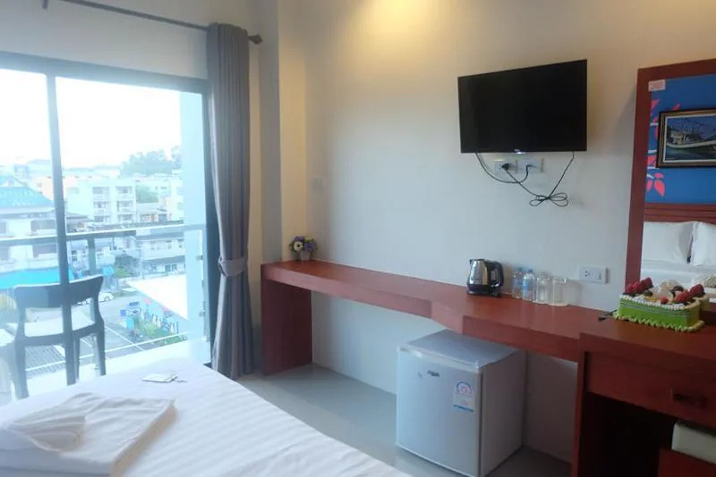 A hotel room with a bed, a desk with a television, a mini-fridge, and a view of a swimming pool and buildings.