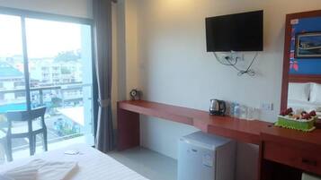 A hotel room with a bed, a desk with a television, a mini-fridge, and a view of a swimming pool and buildings.