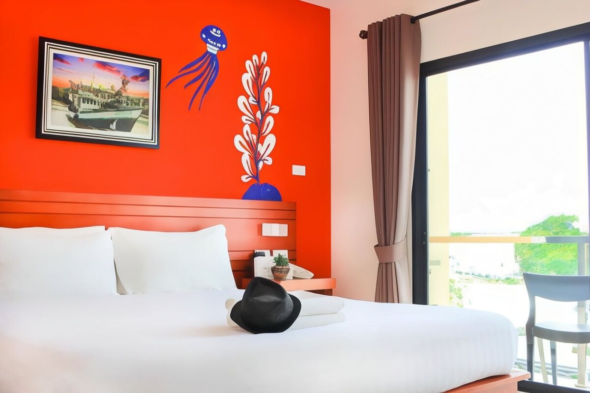 Habitación de hotel con cama, un cuadro en la pared, planta en maceta y vistas a través de la ventana.