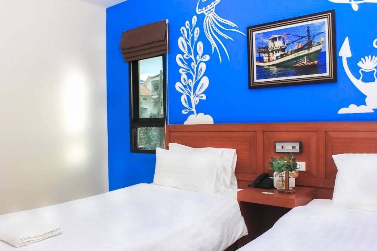 A hotel room with two beds, a window, a wall mural, a framed picture of a boat, and a nightstand with a phone and a plant.