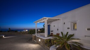 A white building with a stone base, a covered porch, and a palm tree in the foreground. - Stavroula Studios (Mykonos)
