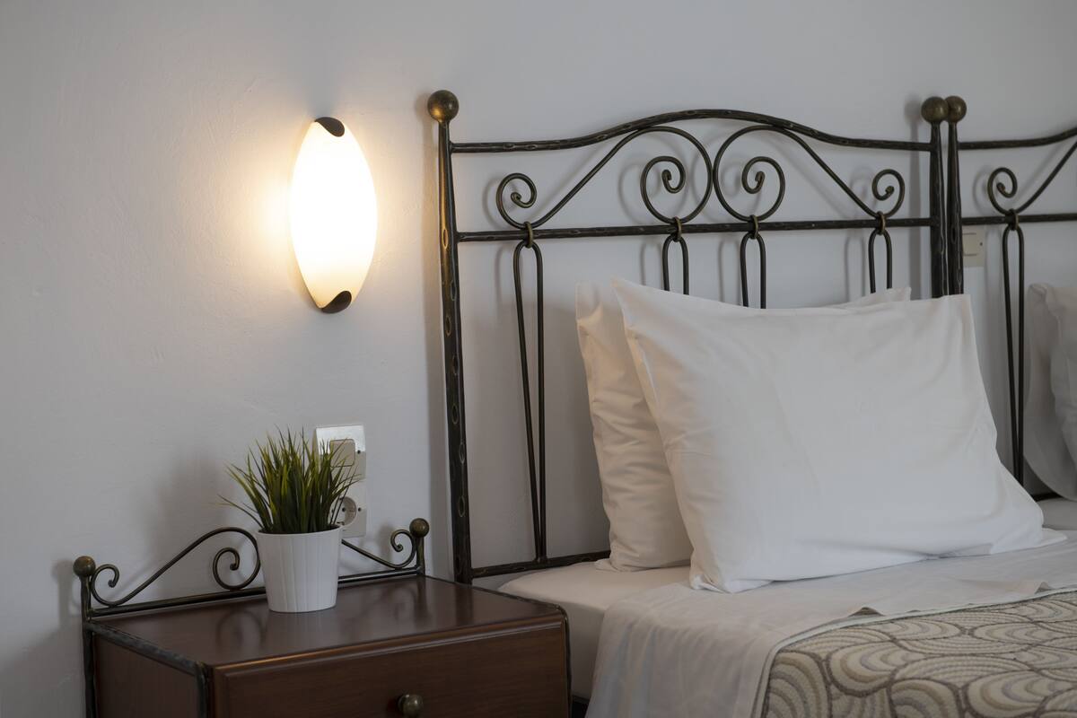 A neatly made bed with white bedding, a bedside table with a potted plant, and a wall-mounted light fixture.