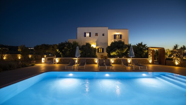 A swimming pool with lounge chairs and umbrellas, surrounded by a landscaped garden and a building with illuminated windows. - Stavroula Studios (Mykonos)