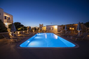 A swimming pool with lounge chairs and umbrellas, surrounded by buildings with warm lighting. - Stavroula Studios (Mykonos)