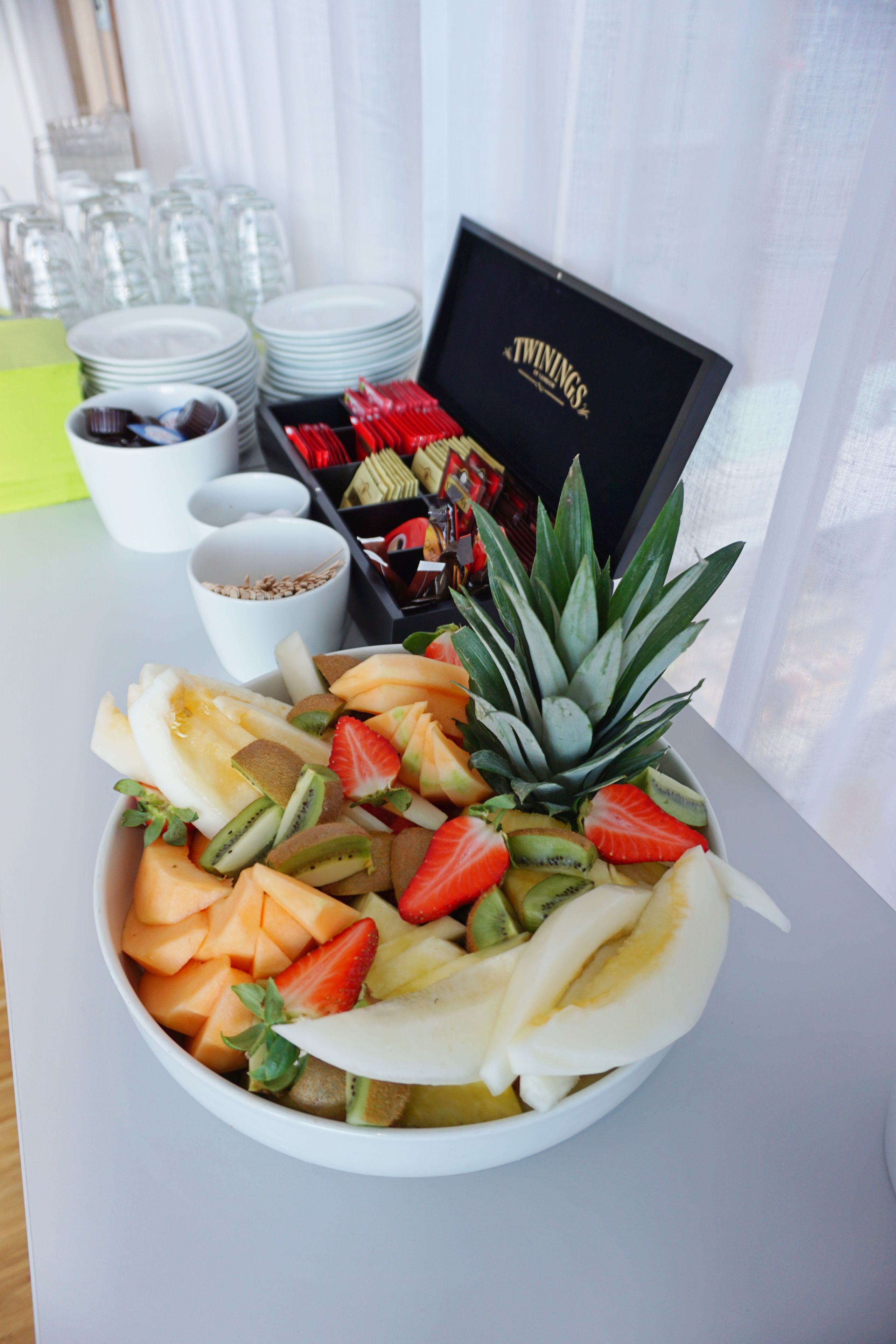 A fruit bowl on a table with a tray of snacks and a box labeled "TWININGS" in the background.