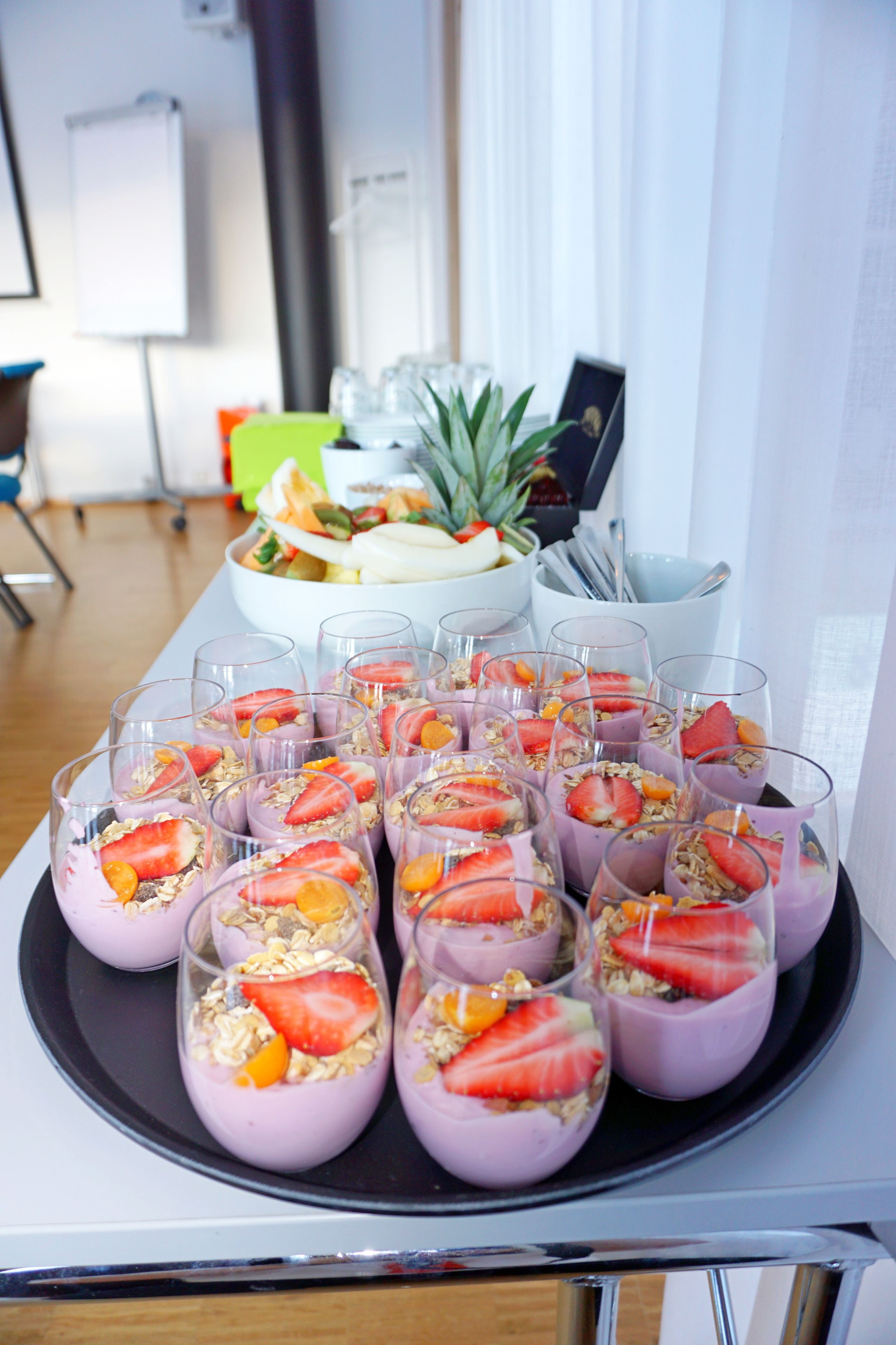 Una bandeja con parafaits, fresas y granola, un tazón de ensalada de frutas y una piña al fondo.