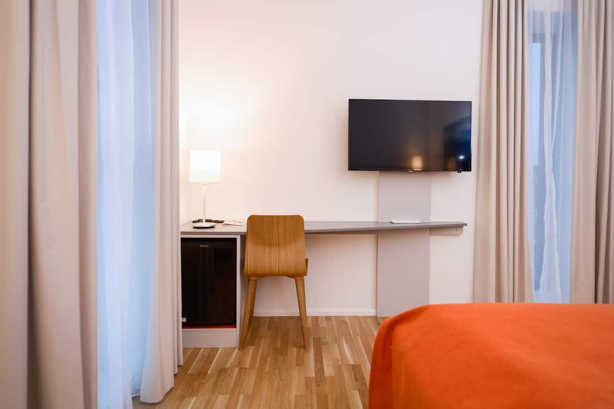 A hotel room with a bed, a desk with a chair, a television mounted on the wall, and a small kitchenette.