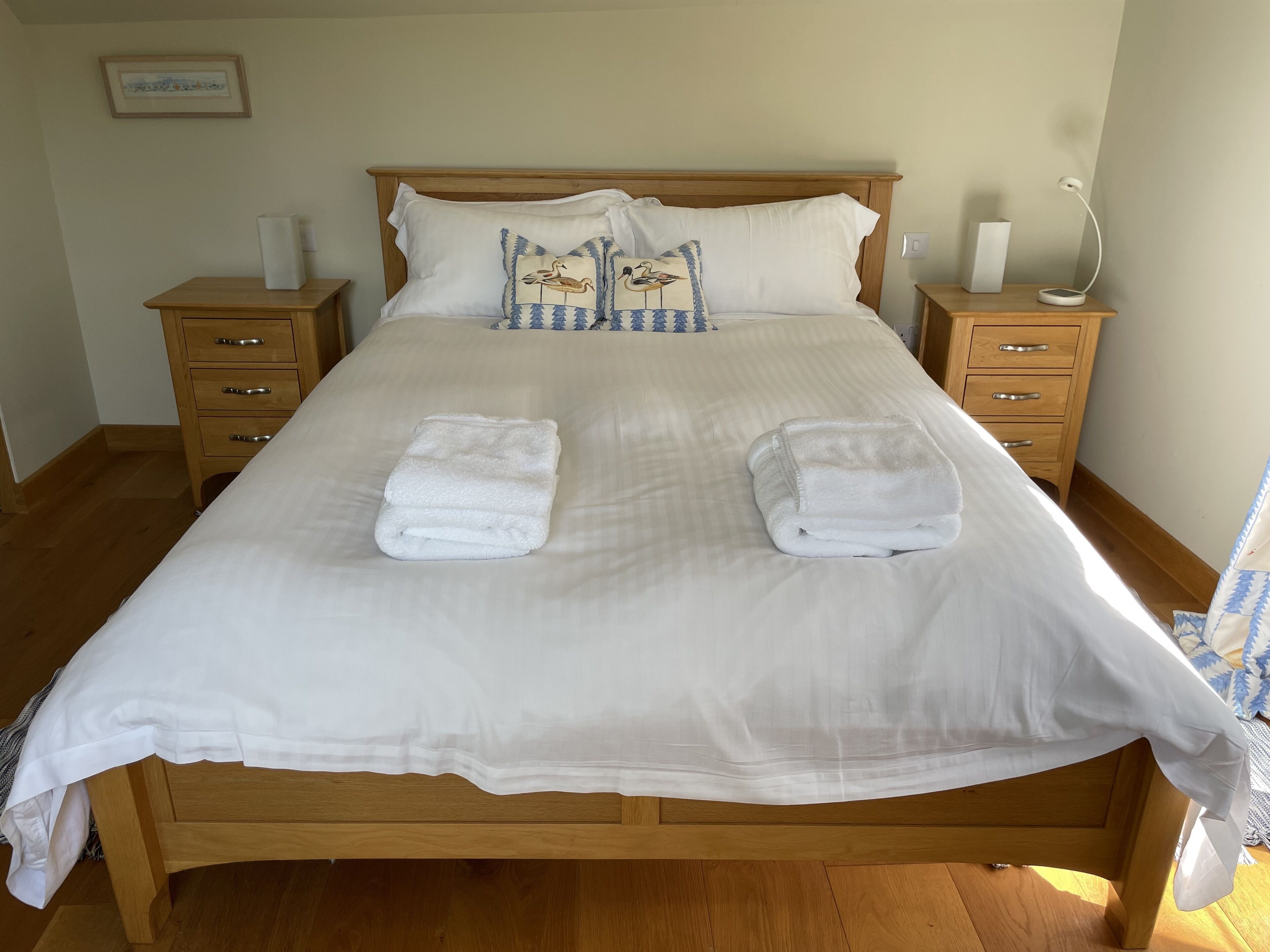 A neatly made bed with two folded towels, wooden headboard, bedside tables with lamps, and a framed picture on the wall.