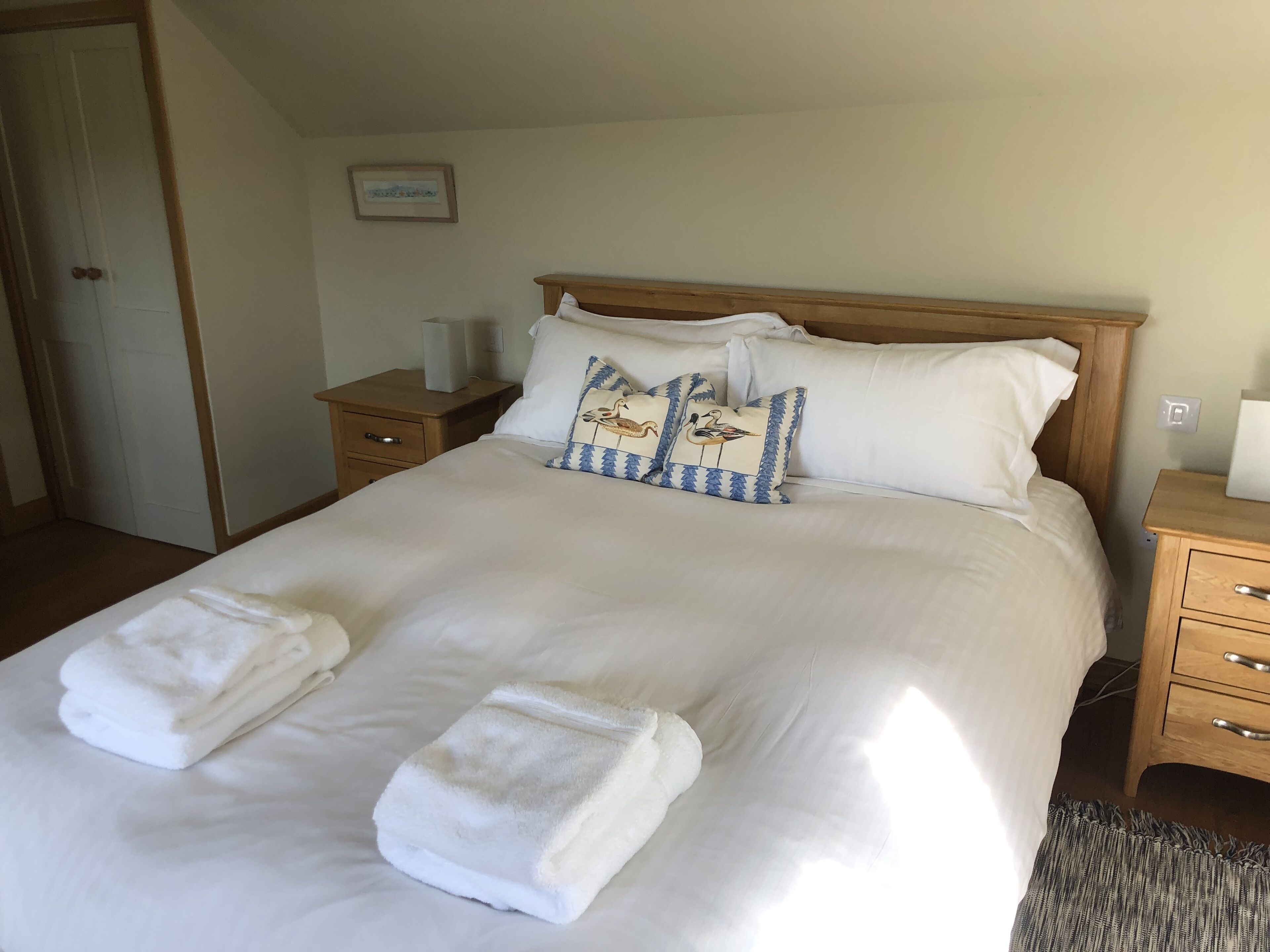 A neatly made bed with white linens and two towels, a wooden headboard, and a bedside table with a lamp.