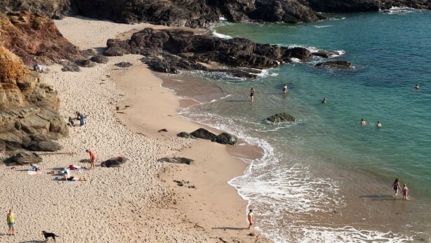 Ein Strand mit felsigen Klippen, klarem Wasser und Menschen, die schwimmen und am Ufer entlanggehen.