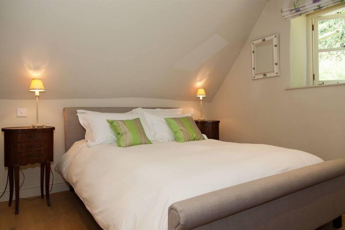 A neatly made bed with white bedding and green pillows, two bedside tables with lamps, a window with a view of greenery, and a small decorative mirror on the wall.