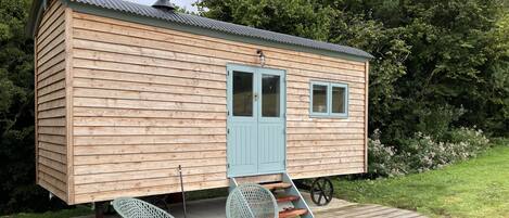 A small wooden cabin with a blue door and windows, a wheelbarrow, and two chairs on a wooden deck.