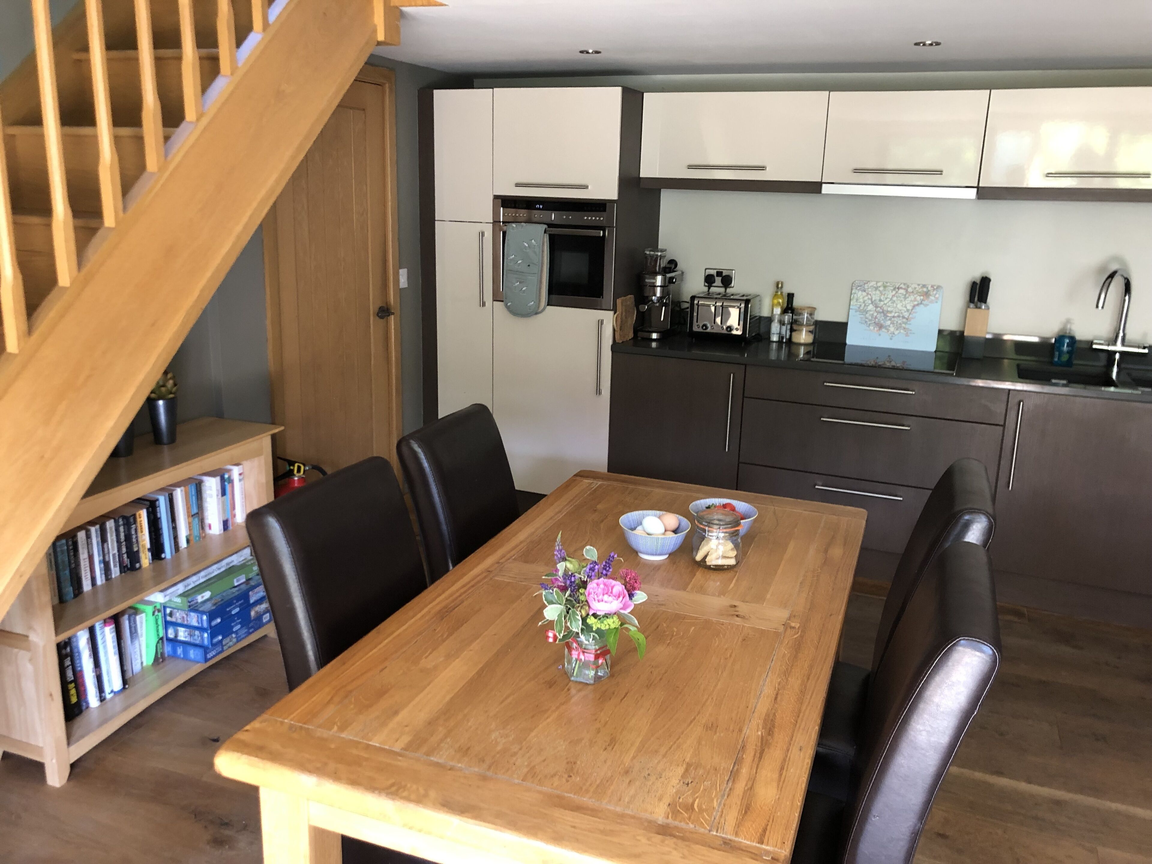 A modern kitchen with white cabinets, a wooden dining table, and a staircase.