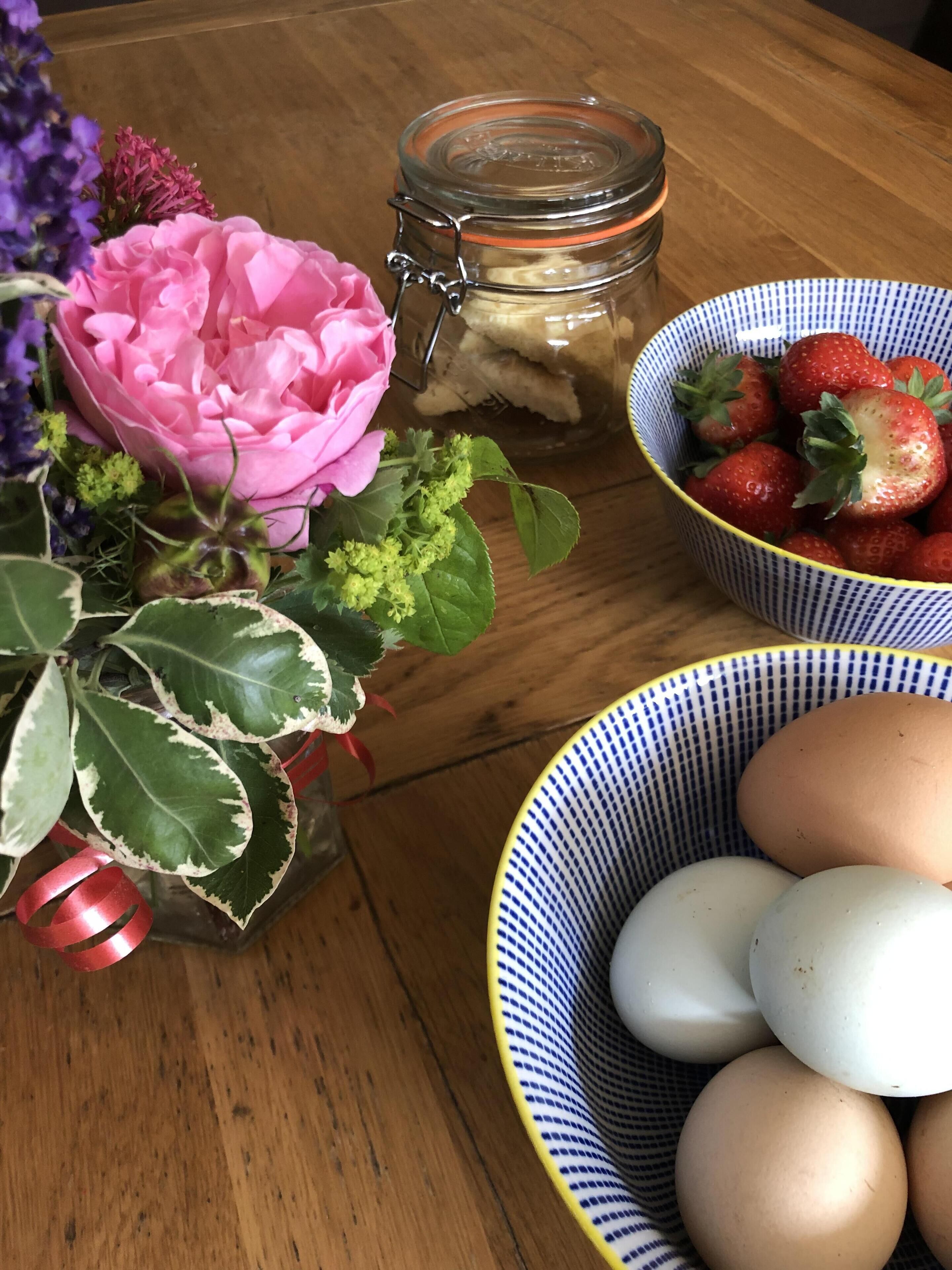 Ett träbord med ett glasburk, en skål med jordgubbar, en skål med ägg och en rosa blomsteruppsättning.