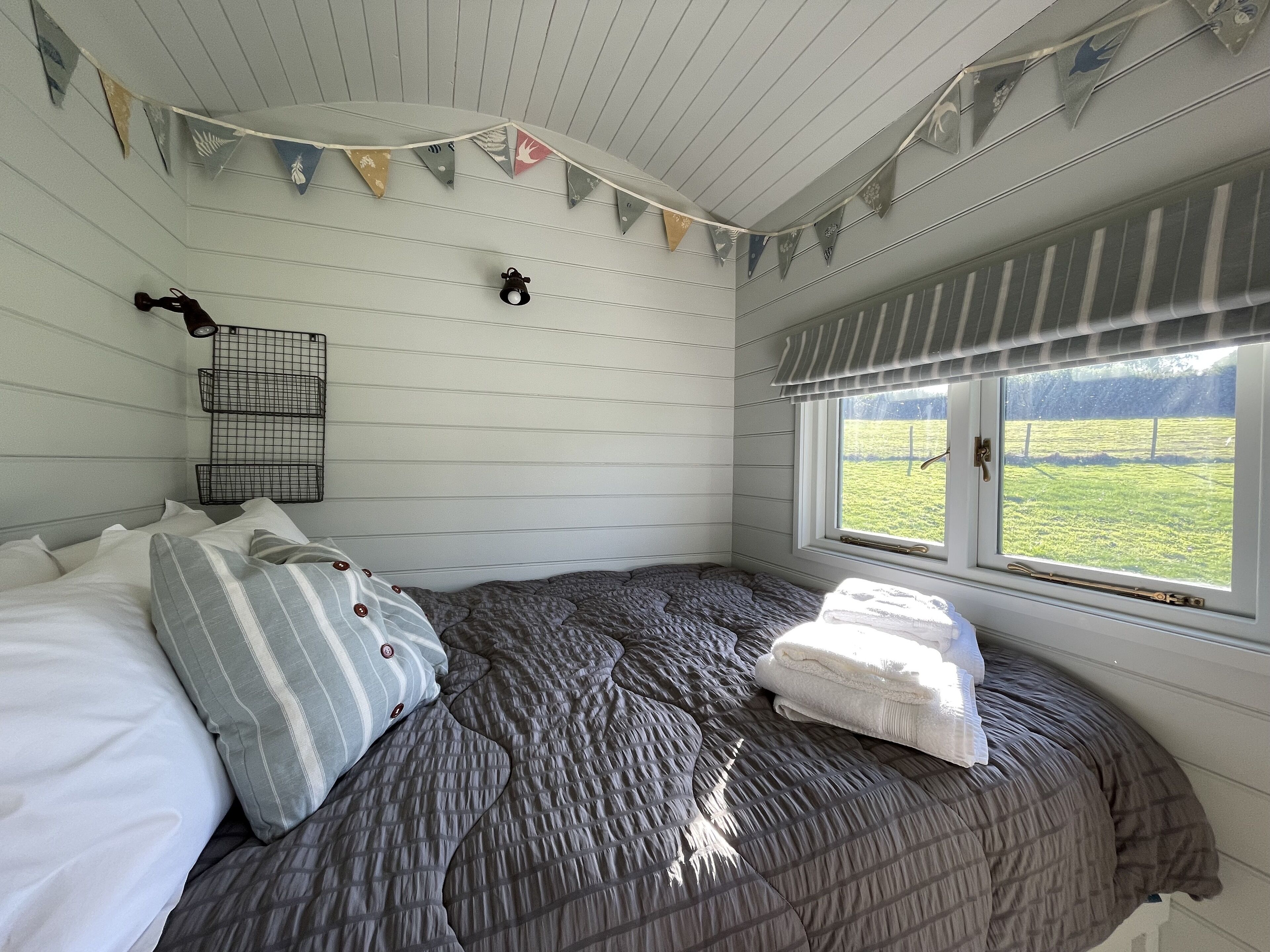 Un dormitorio con una cama, almohadas, una ventana con vistas a un campo verde y una guirnalda de banderas triangulares en el techo.