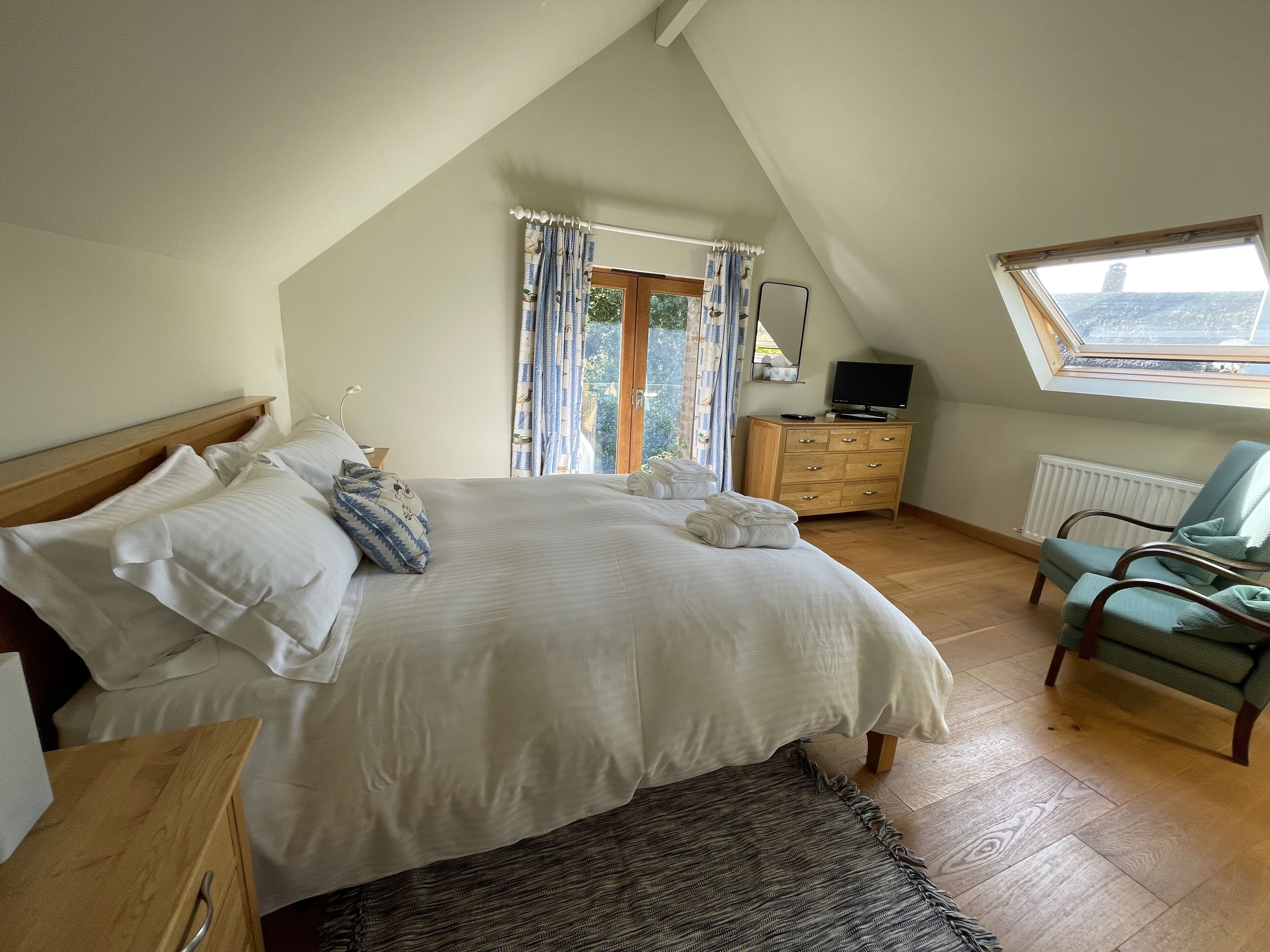 Ein Schlafzimmer mit einem großen Bett, einer Holzkommode, einem grünen Sessel, einem Fenster mit Vorhängen und einem Oberlicht.