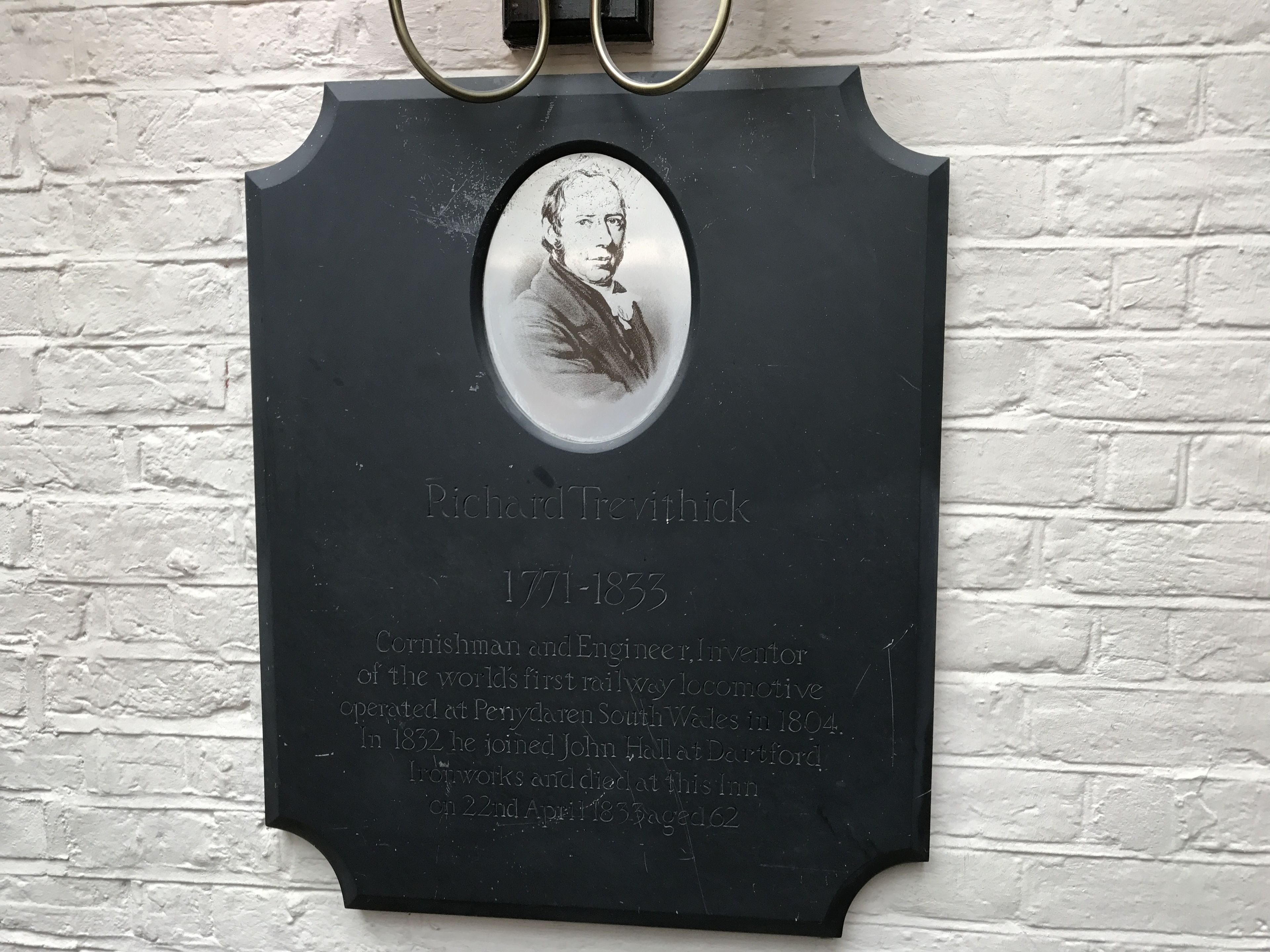 En sort plade med et ovalt portræt og tekst, der beskriver Richard Trevithicks liv og bedrifter, herunder hans rolle som en mand fra Cornwall og ingeniør, samt hans opfindelse af verdens første jernbanelokomotiv.