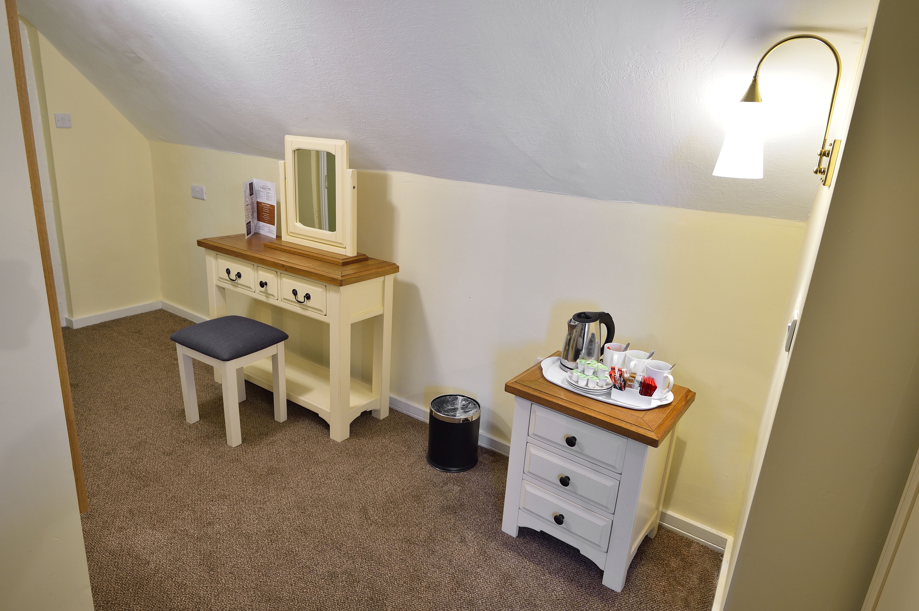 Un dormitorio pequeño y ordenado con una cómoda de madera, un espejo, una mesita con una tetera y tazas, y un cubo de basura.
