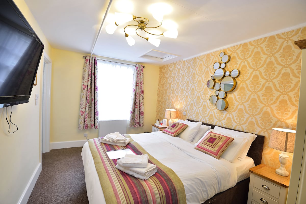 A hotel room with a bed, bedside tables, a TV, a window with curtains, and a patterned wall.