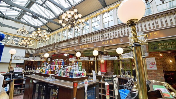 An interior of a bar with multiple beer taps, a counter with various bottles, and a high ceiling with chandeliers. - The Royal Victoria & Bull Hotel (Dartford)