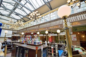 L’intérieur d’un bar, avec de nombreux robinets à bière, un comptoir où sont exposées diverses bouteilles, et un plafond haut orné de lustres.