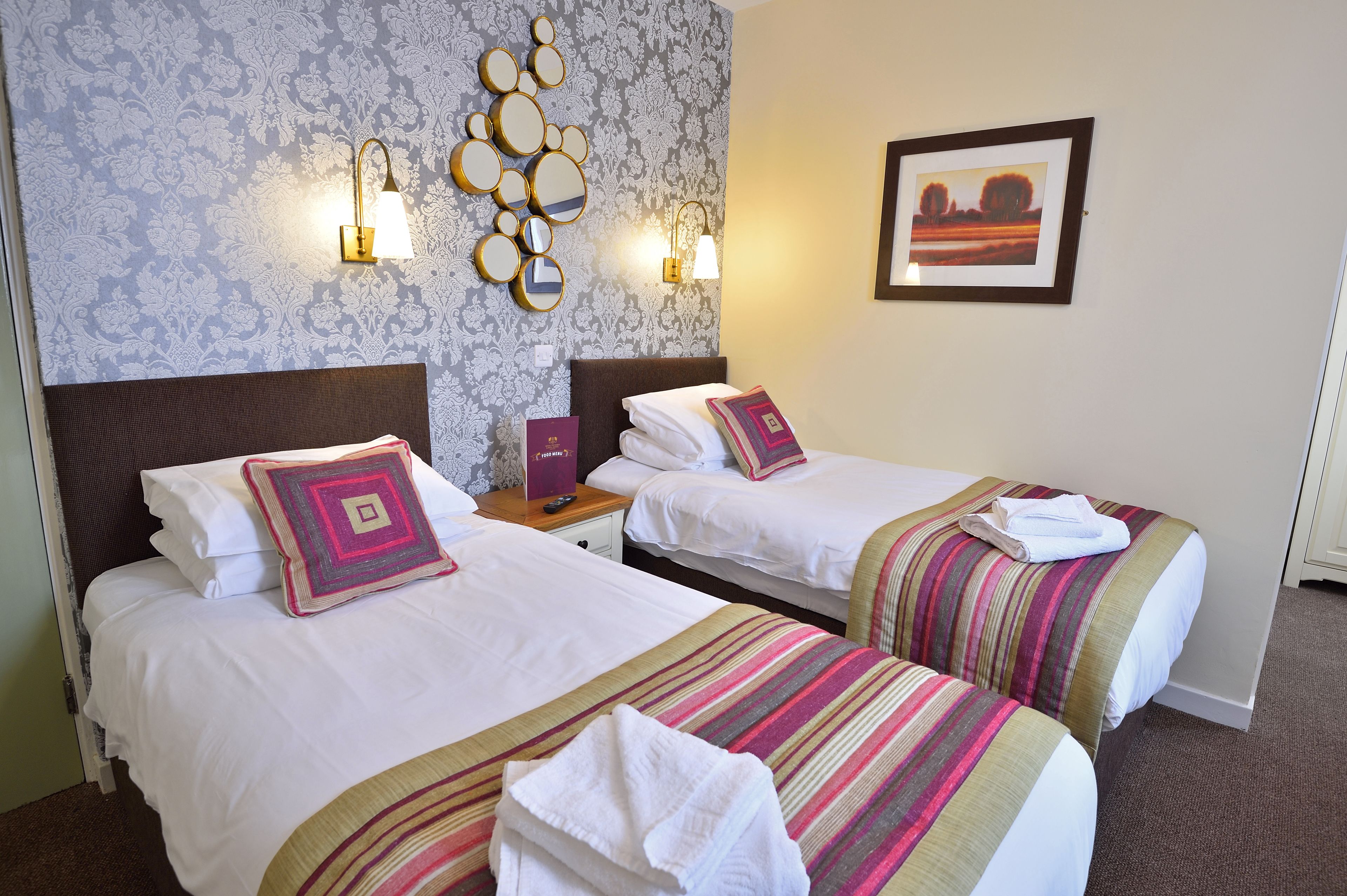 A hotel room with two beds, a patterned wallpaper, wall sconces, a framed picture, and a bedside table with a remote control.