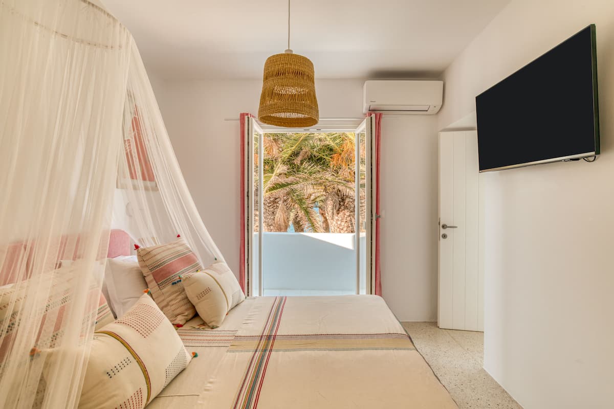 Un dormitorio con cama, lámpara colgante, televisor en la pared y puerta que da a un balcón con vistas a palmeras.