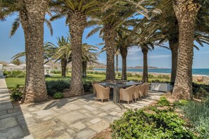 A patio with a round table and chairs, surrounded by palm trees and a view of the sea. - Villa Bellonia (Paros)