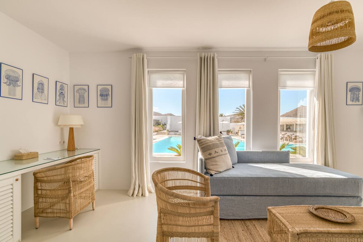 Una habitación con escritorio de vidrio, muebles de mimbre, sofá azul y ventanas grandes con vistas a una piscina y edificios.