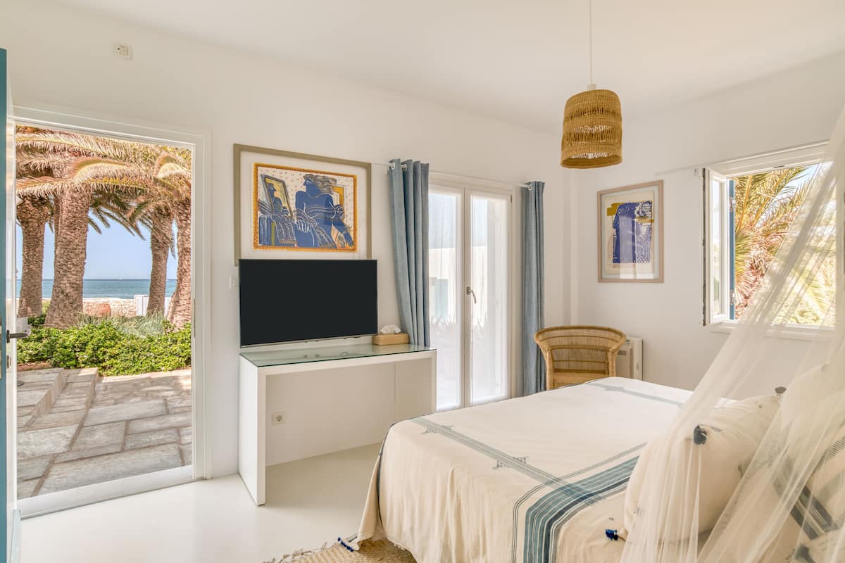 Un dormitorio con cama, televisor sobre un mueble de vidrio y vistas a palmeras y al mar.
