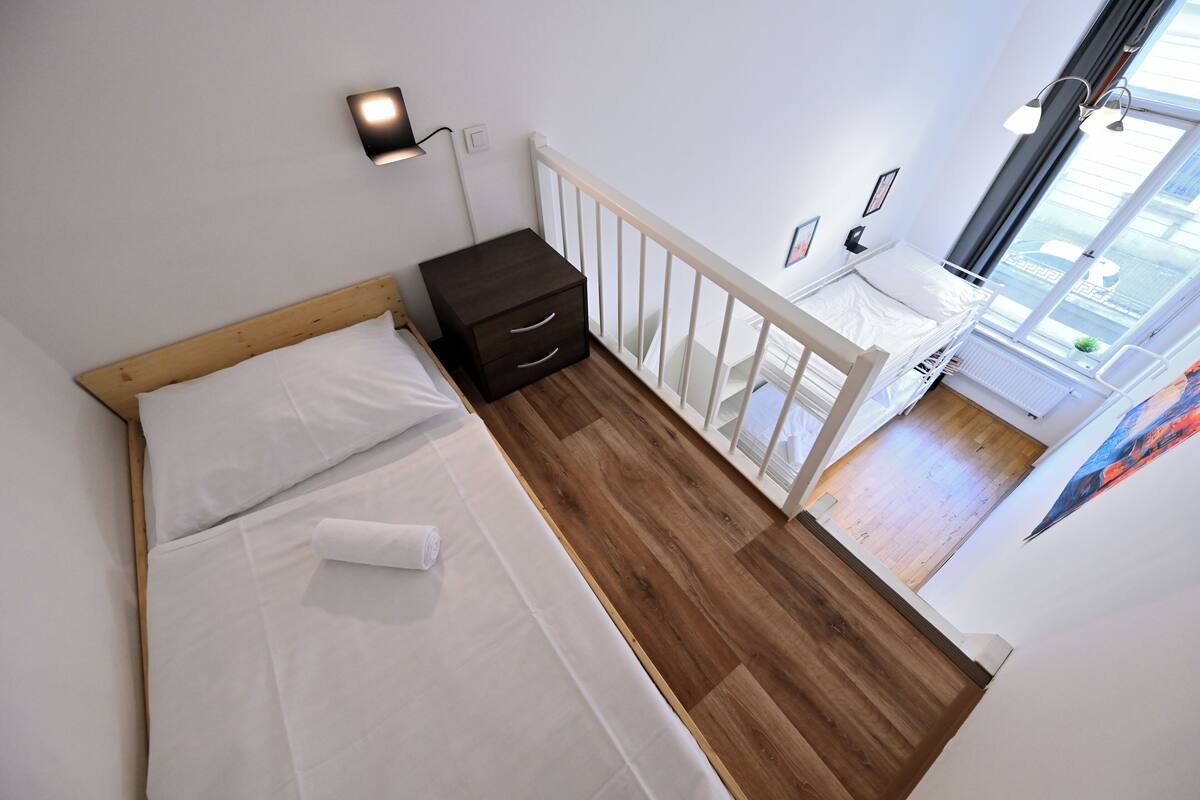 A bedroom with a wooden floor, a bed with white bedding, a nightstand, and a window with a view of the outside.