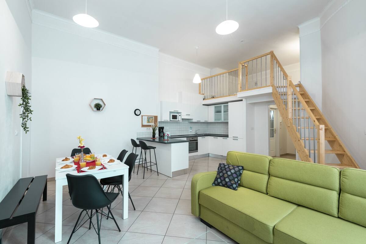A modern kitchen with white cabinets and a central island, a dining area with a white table and black chairs, and a green sofa with a patterned pillow.