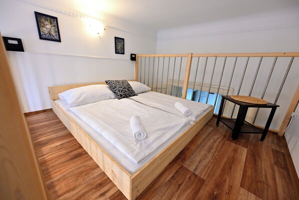 A wooden bed with white linens and two rolled towels, a small table with a woven top, and a framed picture on the wall. - Hybernska Apartments (Prague)