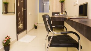 Une chambre d’hôtel moderne avec une porte en bois foncé, une chaise noire, un bureau avec un ordinateur portable, une petite plante en pot et un miroir avec des lumières.