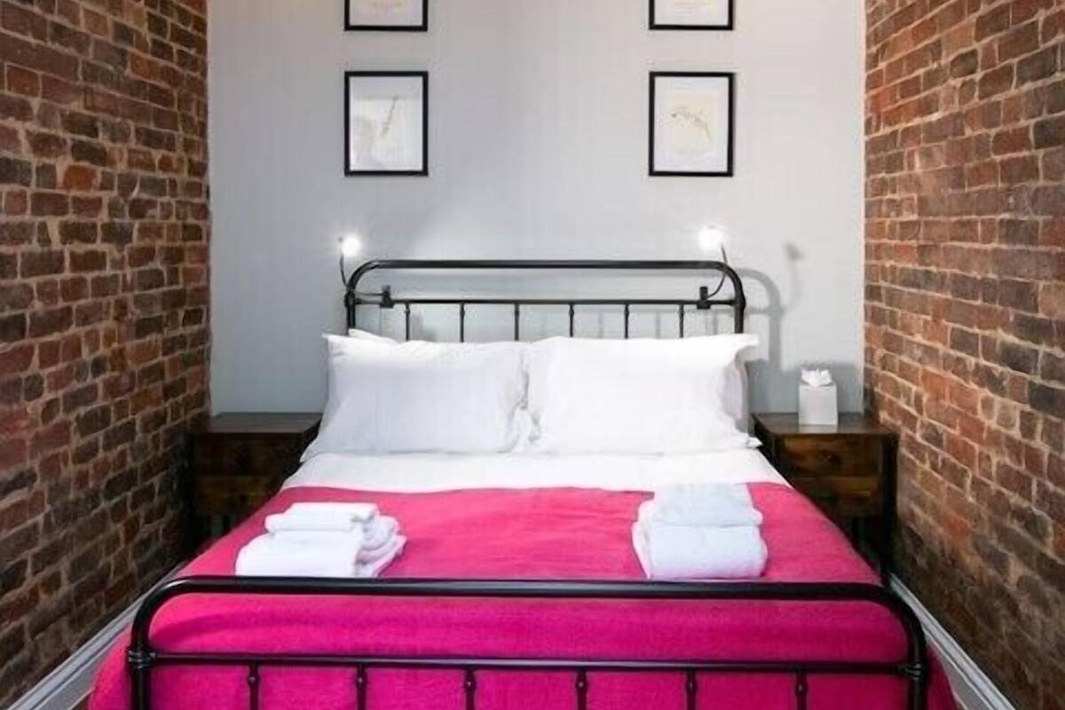 Ein Zimmer mit Ziegelwand, einem Metallbettgestell, weißer Bettwäsche und einer rosa Decke. An der Wand hängen drei gerahmte Bilder und ein Nachttisch mit einer Lampe.