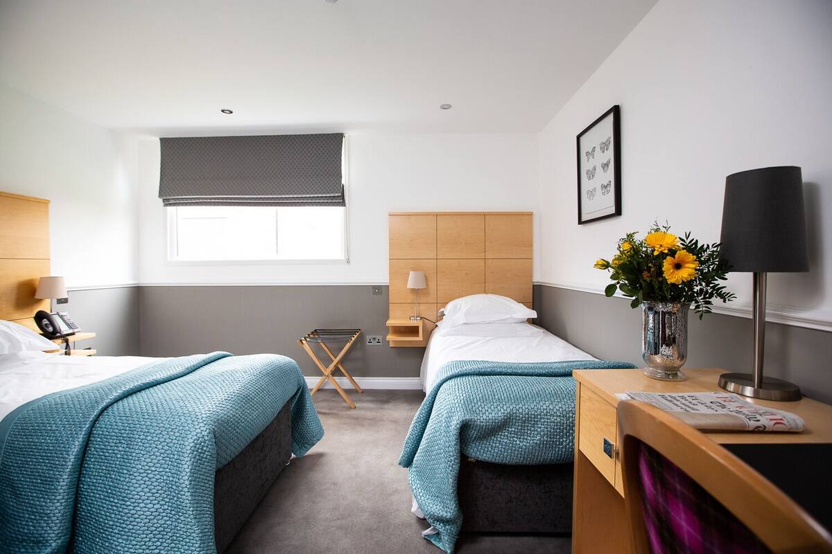 Ein Hotelzimmer mit zwei Betten, einem Fenster mit Jalousie, einem Nachttisch mit Lampe, einem kleinen Tisch mit Blumenvase und einem Holzkopfstück.