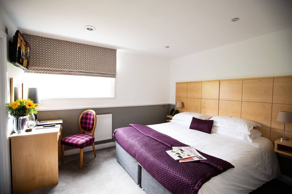 Ein Hotelzimmer mit einem Bett, einem Sessel, einem Schreibtisch, einer Heizung, einem Fenster mit Jalousien und einer Wanduhr.