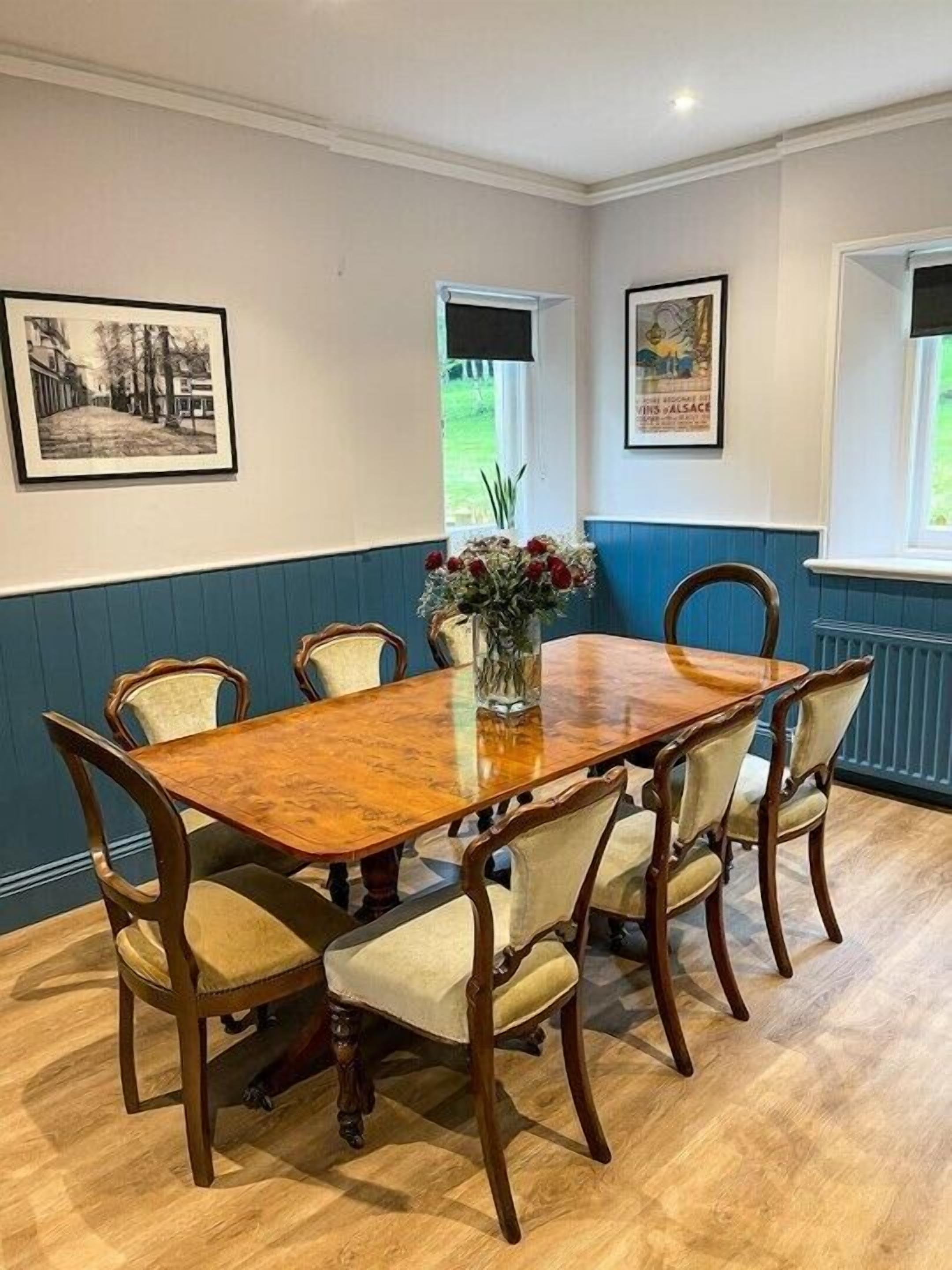 A dining room with a wooden table and six chairs, a vase with flowers, and two framed pictures on the walls.