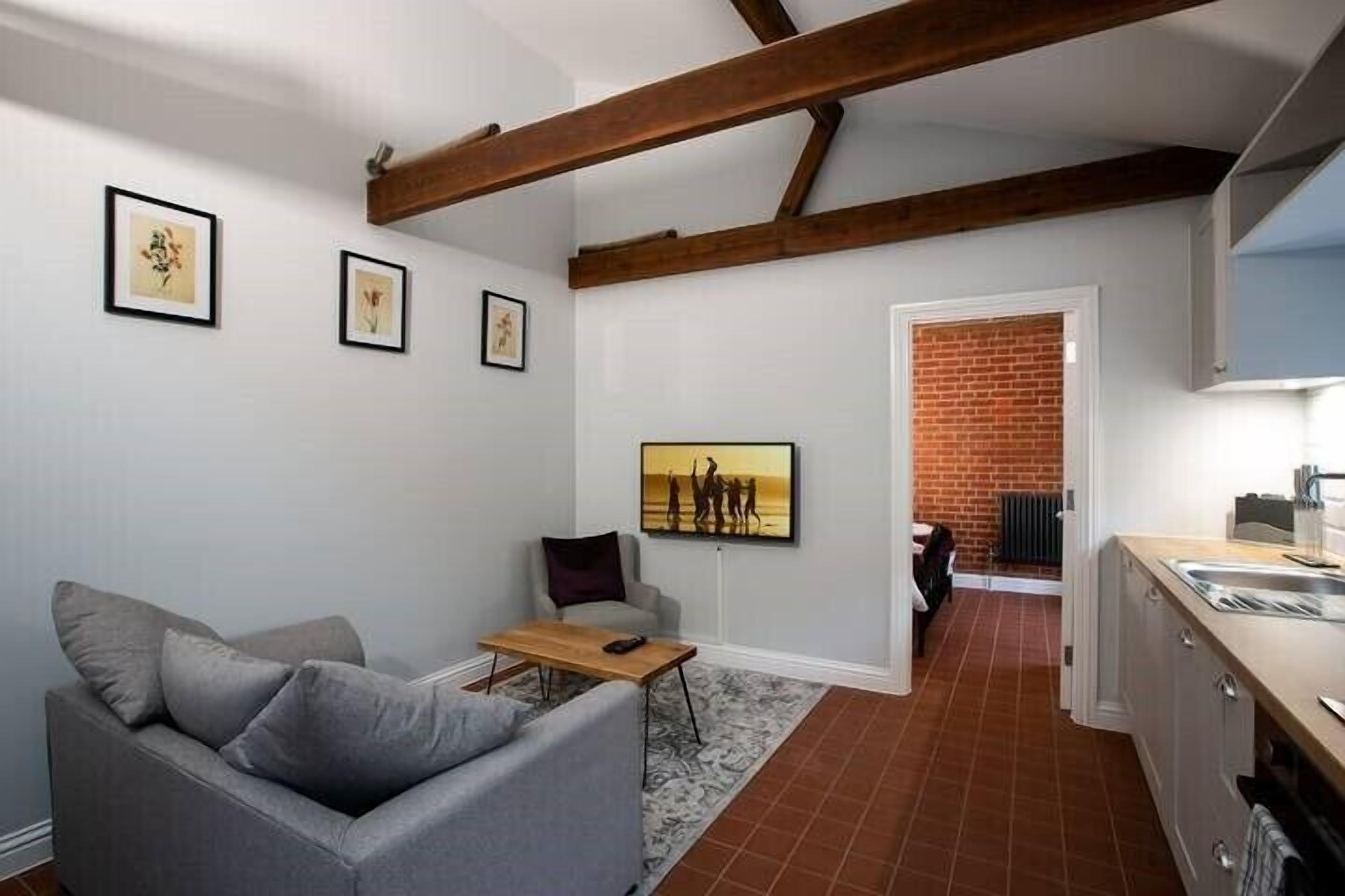 Ein kompakter Wohnraum mit grauem Sofa, kleinem Tisch und Küchenzeile mit Backsteinwand und weißen Schränken.
