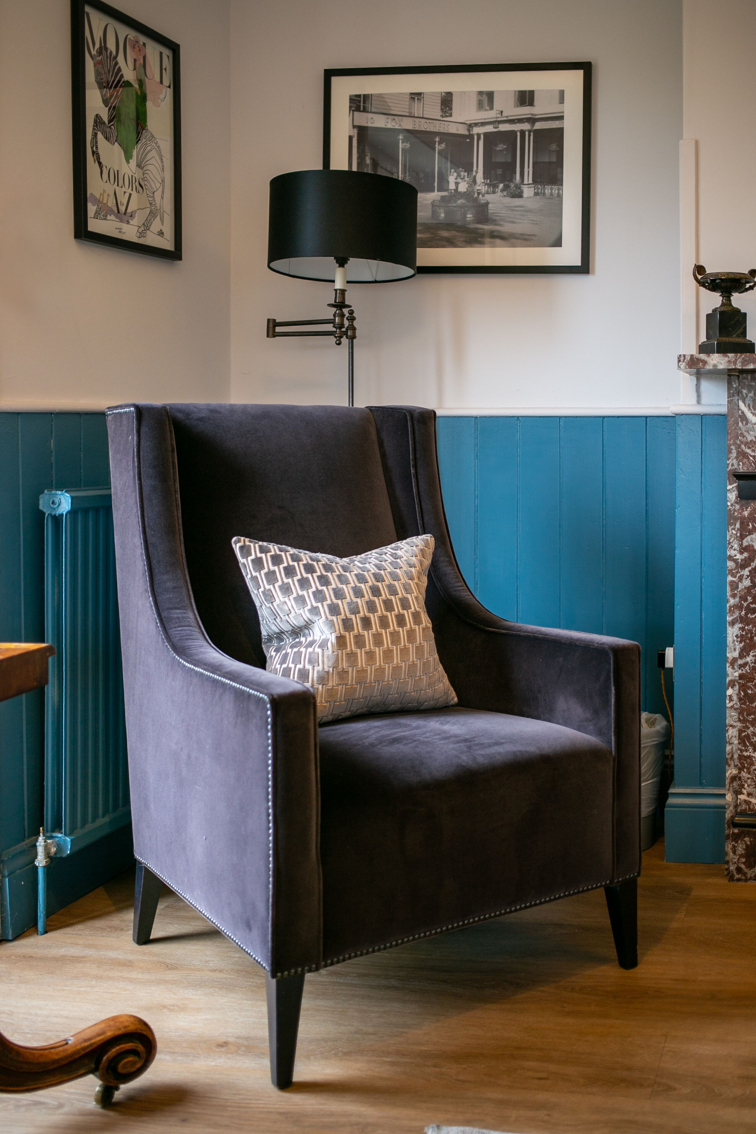 A dark-colored armchair with a patterned cushion in a room with teal walls, a black floor lamp, and framed pictures on the wall.