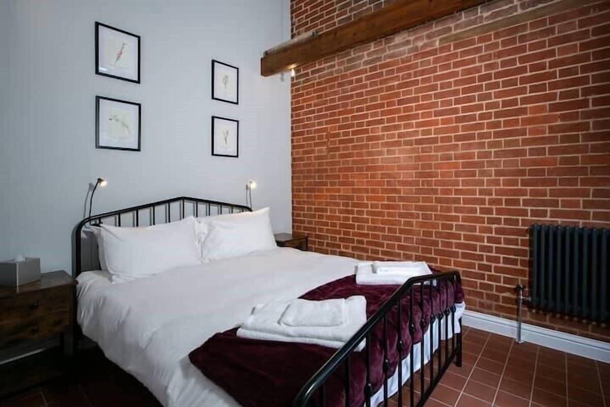 Ein ordentlich bezogenes Bett mit weißen Bettwäsche und einer roten Tagesdecke, eine Backsteinwand und eine Heizung.