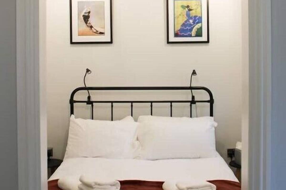 Ein ordentlich bezogenes Bett mit weißen Leinen und einem braunen Kopfteil, zwei gerahmte Kunstwerke an der Wand und ein schlichter Nachttisch.