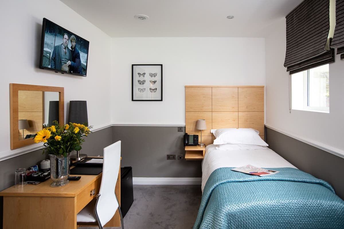 Ein Hotelzimmer mit Bett, Schreibtisch mit Stuhl, Fernseher, einem gerahmten Bild und einem Fenster mit Jalousien.