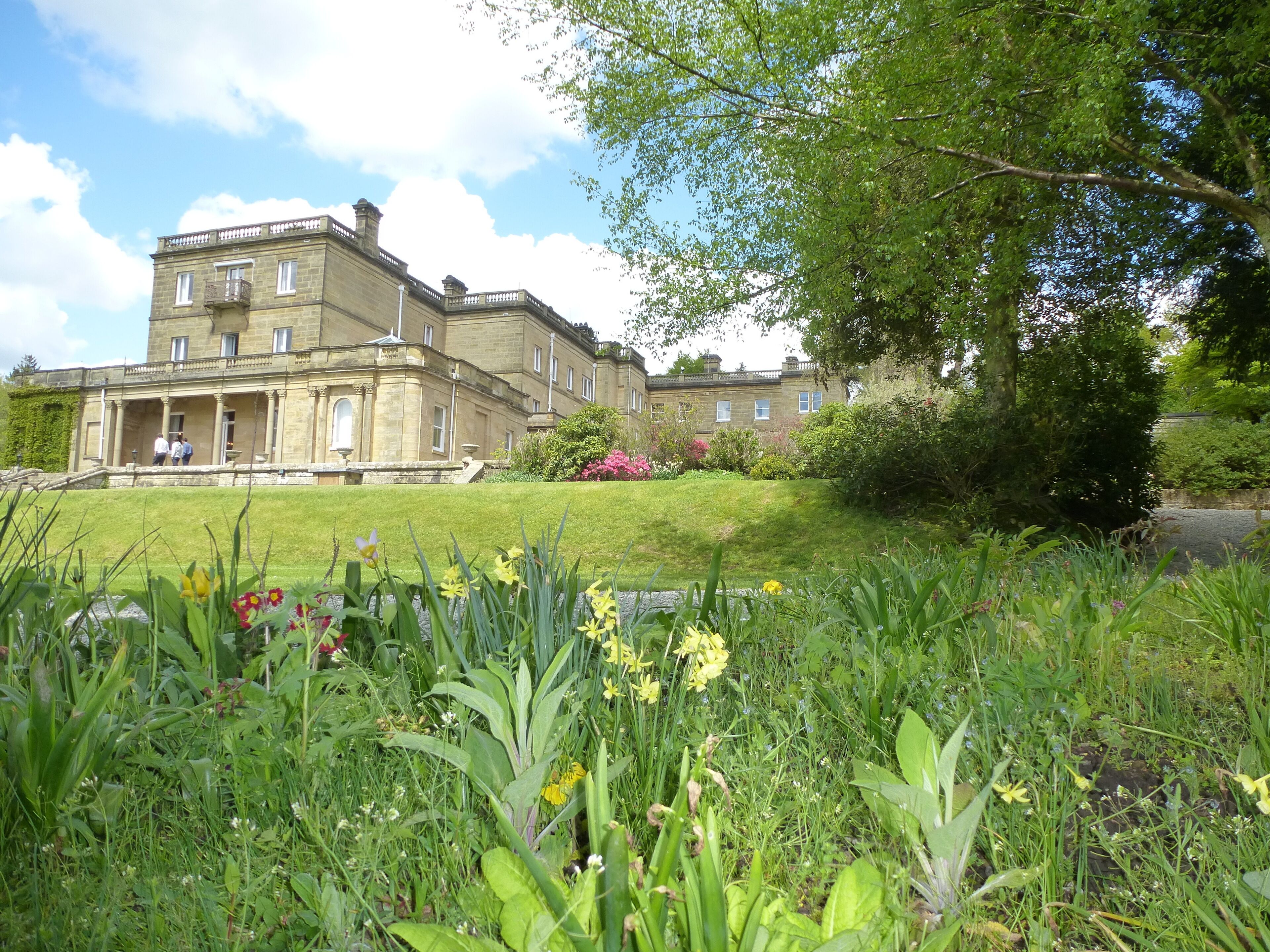 A large, historic building with a symmetrical facade, a central entrance with columns, and a well-maintained garden with flowering plants and a lawn.