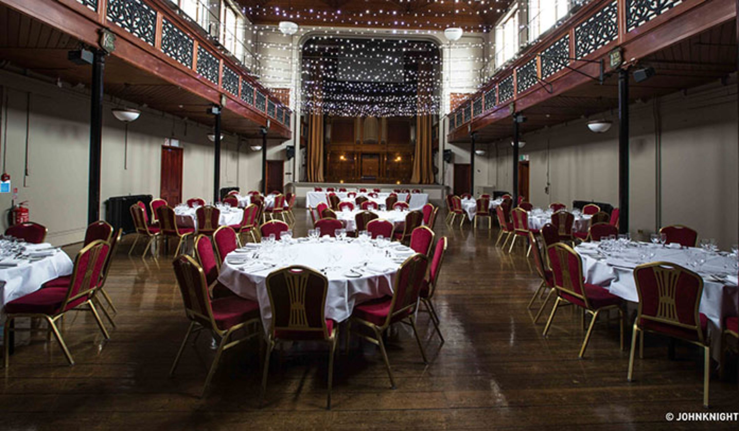 A banquet hall with round tables and red chairs, wooden floors, and a high ceiling with decorative lighting.