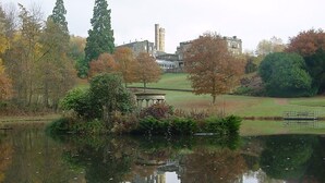 A serene lake with a gazebo, surrounded by trees and a historic building with a tower in the background. - Accommodation at Salomons Estate (Royal Tunbridge Wells)